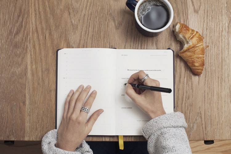 a shot of a hand writing in a notepad with coffee next to it on a table